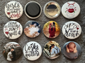 Collection of various 1.5 inch round pin buttons and magnets with different designs.  The buttons and magnets are photographed against a soft gray surface background.