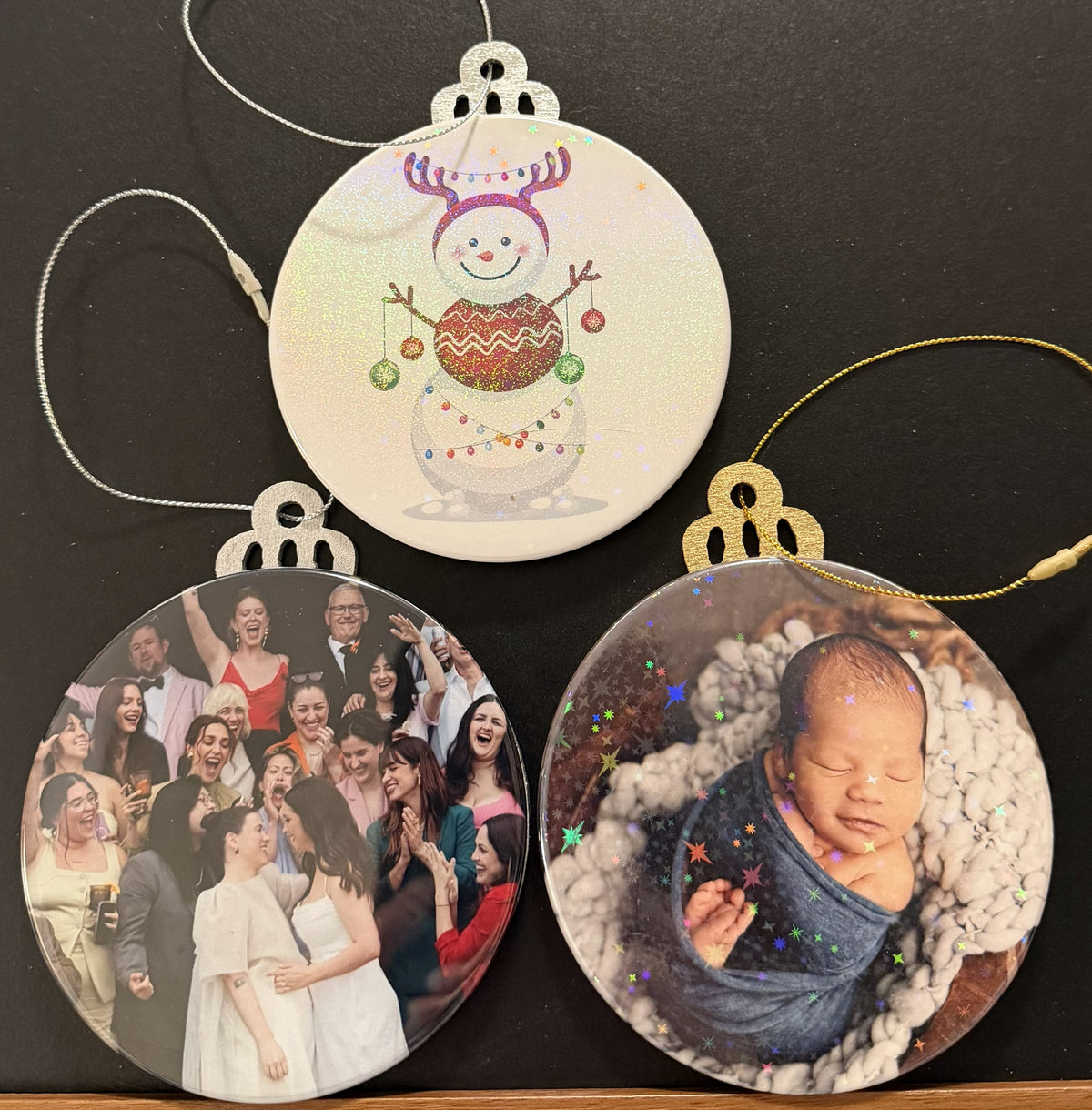 Three samples of Holiday/Christmas ornaments featuring a snowman and two babies on a dark surface.  Gold or silver colored wooden ornament backing with a gold or silver string to hang the ornament.  One ornament has a clear covering, while the other  have clear holographic covering.