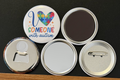 A white round button with 'I love someone with autism in multi-colored text, with yellow stars
and a multi-colored puzzle piece heart on a white background.  the other 4 buttons show the backside options which include magnet magnetic bottle cap opener, pin back button and pocket mirror.  Photographed on a black background.
