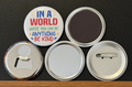 A white round button with 'In a world where you can be anything, be kind' in multi-colored text, with yellow and blue puzzle pieces on a white background.  The other 4 buttons show the backside options which include magnet magnetic bottle cap opener, pin back button and pocket mirror.  Photographed on a black background.