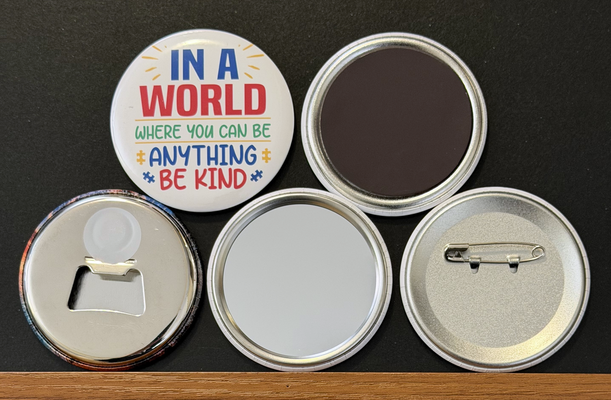 A white round button with 'In a world where you can be anything, be kind' in multi-colored text, with yellow and blue puzzle pieces on a white background.  The other 4 buttons show the backside options which include magnet magnetic bottle cap opener, pin back button and pocket mirror.  Photographed on a black background.