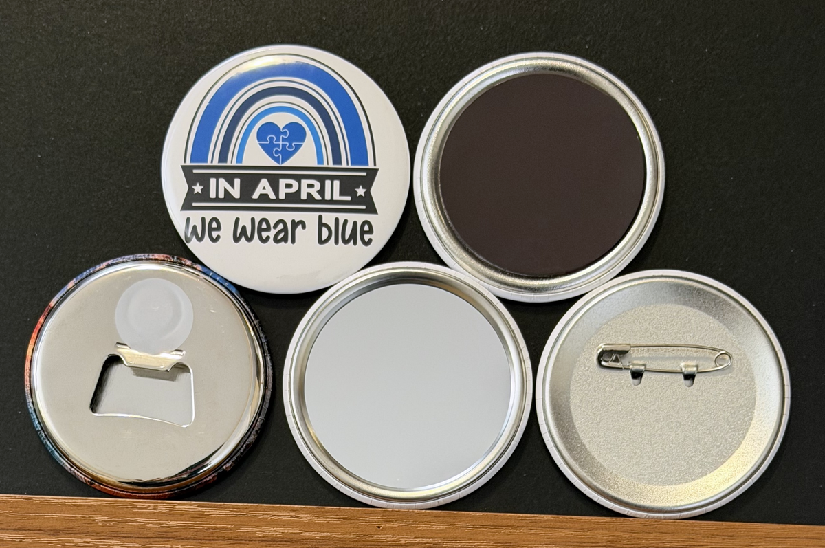 A white round button with 'In April, we wear blue' in black and white text with a blue rainbow and white stars on a white background.  The other 4 buttons show the backside options which include magnet magnetic bottle cap opener, pin back button and pocket mirror.  Photographed on a black background.