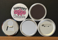 Round white magnet with the message 'Support Squad Cancer' in black and pink text with a pink breast cancer ribbon.  Also in the photo are the four options available for this magnet which include a pin back button, pocket mirror, magnetic bottle opener and magnet.  This image was photographed on a black background.