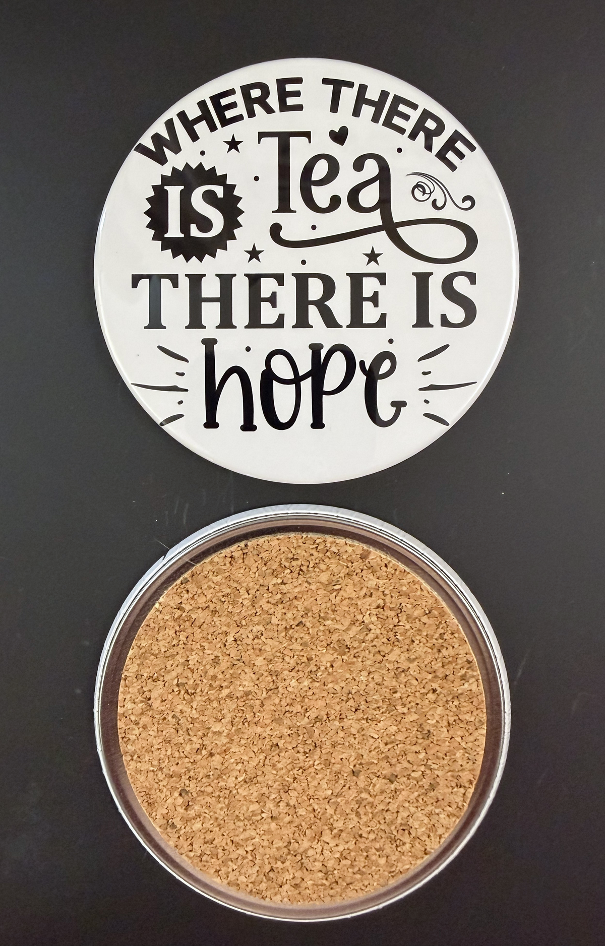 Round white drink coaster with cork backing with 'Where there is tea, there is hope' in black text, small black stars and hearts, photographed on a black background.