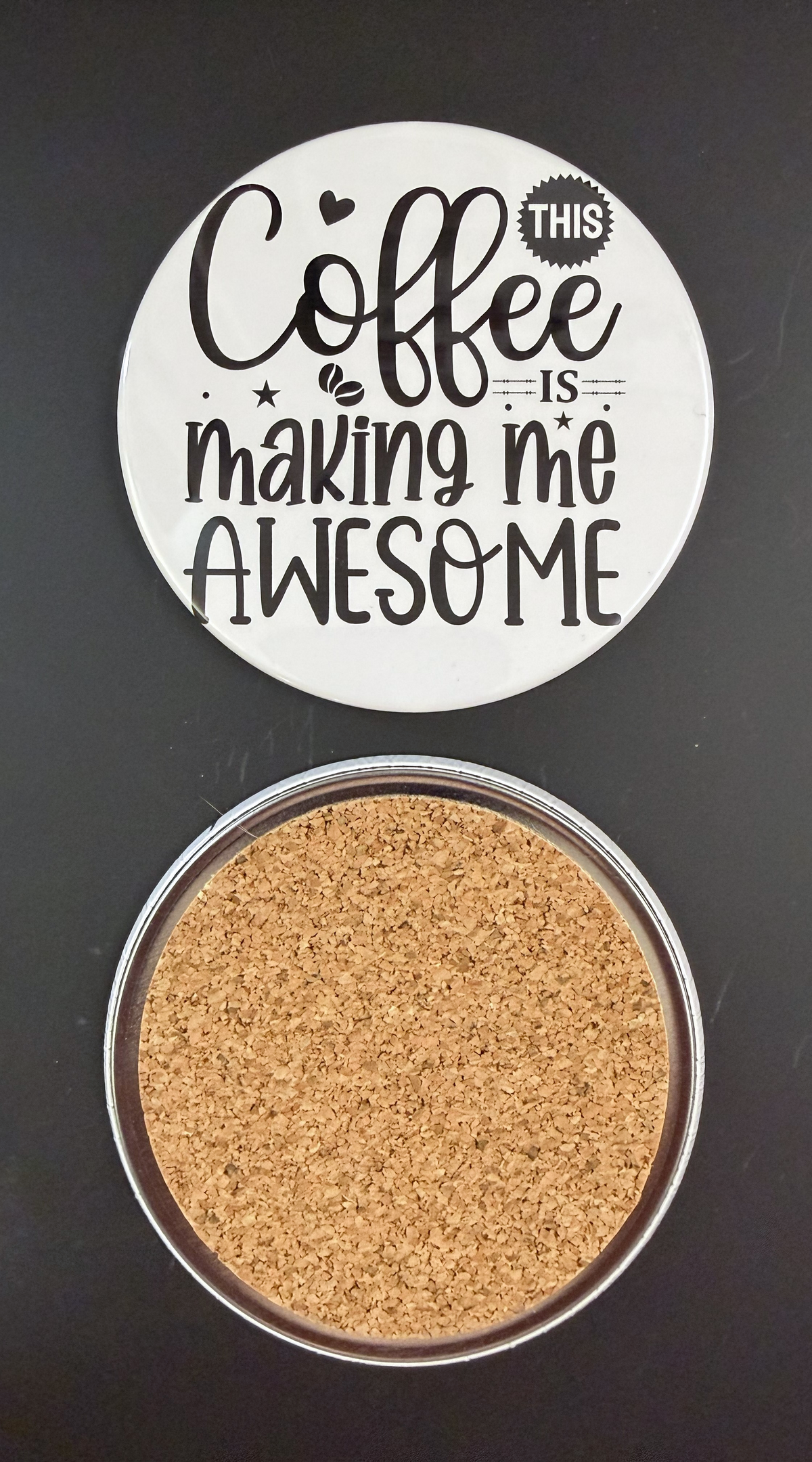 Round white drink coaster with cork backing with 'Coffee is making me awesome' in black text, with little black hearts, stars and coffee beans, photographed on a black background.