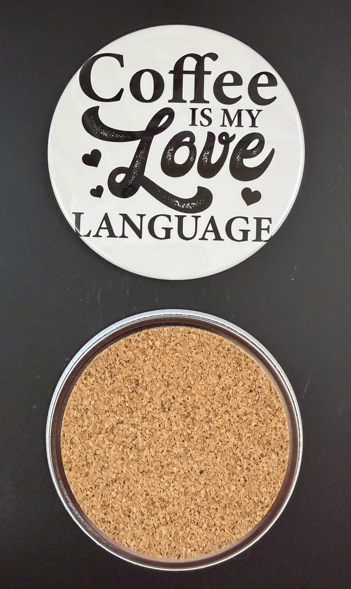 Round white drink coaster with cork backing with 'Coffee is my love language' in black text, with little black hearts, photographed on a black background.