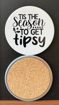 White round drink coaster with a cork backing with black decorative holiday season decor  with text 'Tis the season to get tipsy' on a white background.  This photo was taken on a black background.