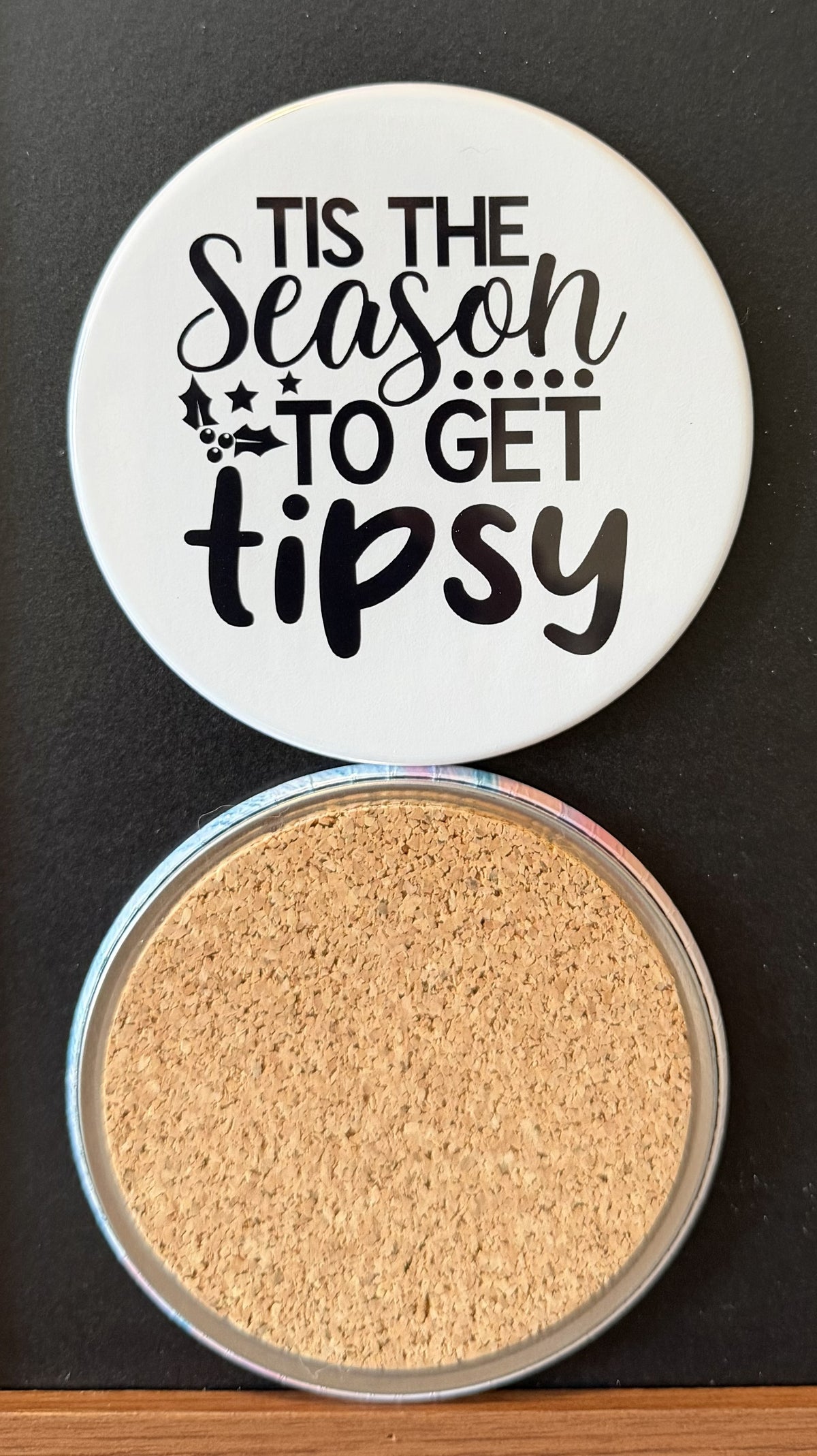 White round drink coaster with a cork backing with black decorative holiday season decor  with text 'Tis the season to get tipsy' on a white background.  This photo was taken on a black background.