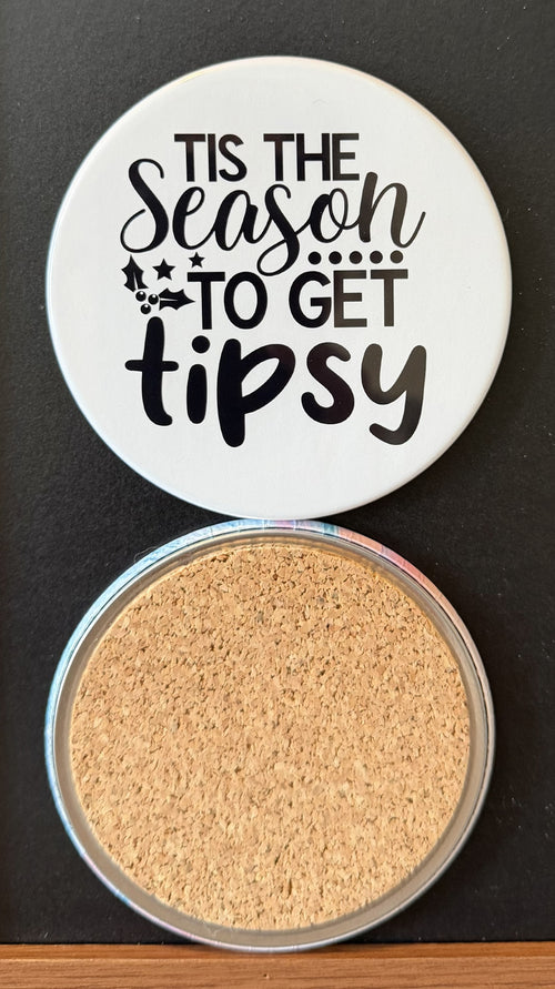 White round drink coaster with a cork backing with black decorative holiday season decor  with text 'Tis the season to get tipsy' on a white background.  This photo was taken on a black background.