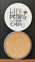 White round drink coaster with a cork backing with black images of a steaming coffee cup and star accents with text 'Life begins with coffee' on a white background.  This photo was taken on a black background.