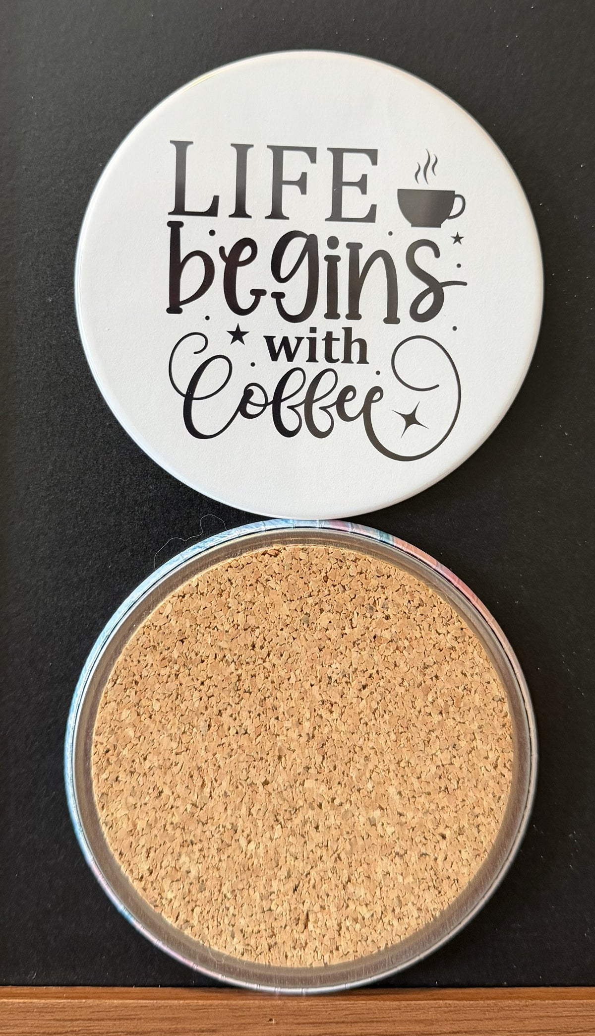 White round drink coaster with a cork backing with black images of a steaming coffee cup and star accents with text 'Life begins with coffee' on a white background.  This photo was taken on a black background.