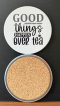 White round drink coaster with a cork backing with a black image of a steaming cup of tea with text 'good things happen over tea' on a white background.  This photo was taken on a black background.