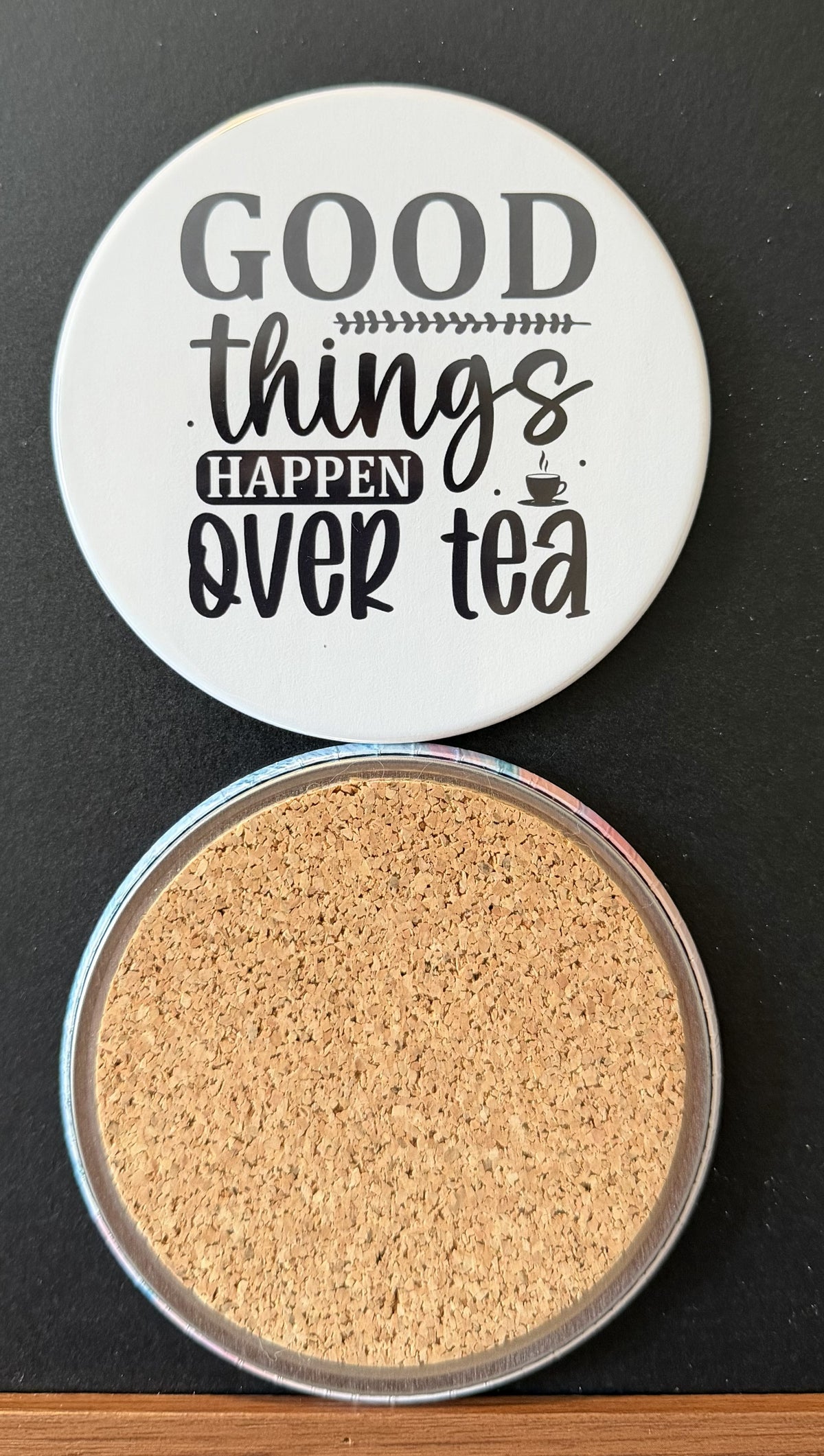 White round drink coaster with a cork backing with a black image of a steaming cup of tea with text 'good things happen over tea' on a white background.  This photo was taken on a black background.