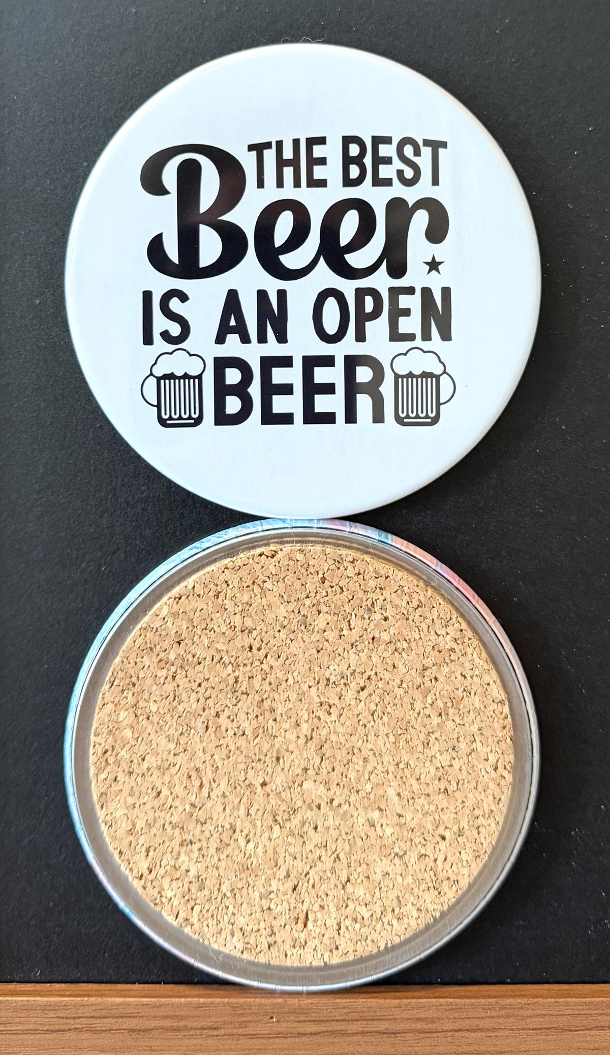 White round drink coaster with a cork backing with two black images of beer mugs with text 'the best beer is  an open beer' on a white background.  This photo was taken on a black background.