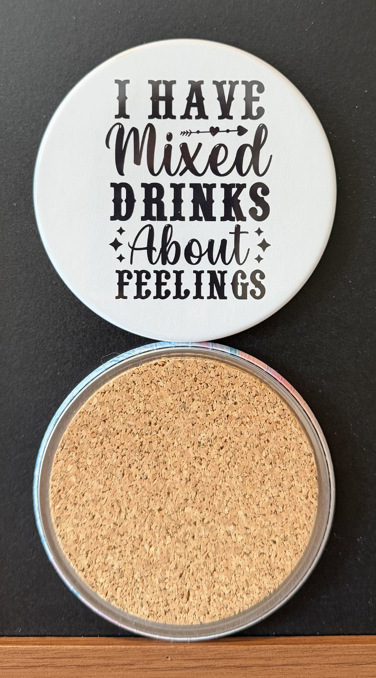 White round drink coaster with a cork backing with black decorative accents with text 'i have mixed drinks about feelings' on a white background.  This photo was taken on a black background.