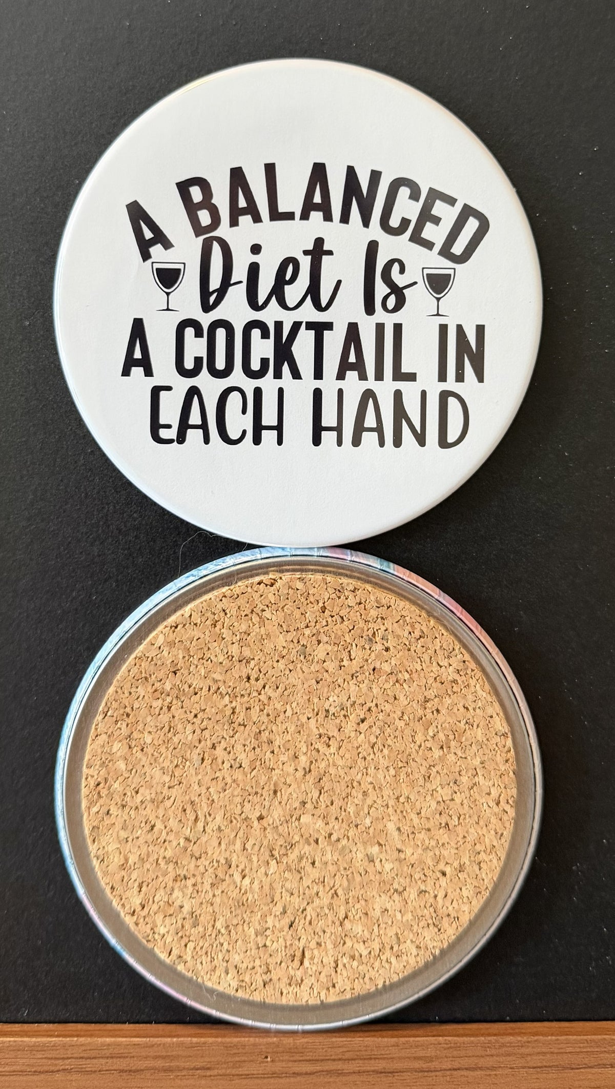 White round drink coaster with a cork backing with 2 black images of cocktail glasses with text 'a balanced diet is a cocktail in each hand' on a white background.  This photo was taken on a black background.