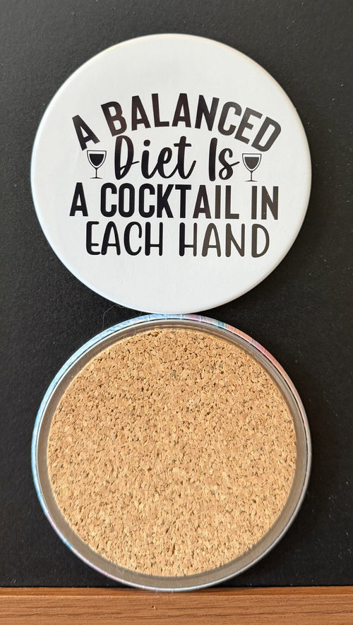 White round drink coaster with a cork backing with 2 black images of cocktail glasses with text 'a balanced diet is a cocktail in each hand' on a white background.  This photo was taken on a black background.