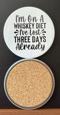 White round drink coaster with a cork backing with black DECORATIVE ACCENTS with text 'I'M ON A WHISKEY DIET, I'VE LOST THREE DAYS ALREADY' on a white background.  This photo was taken on a black background.