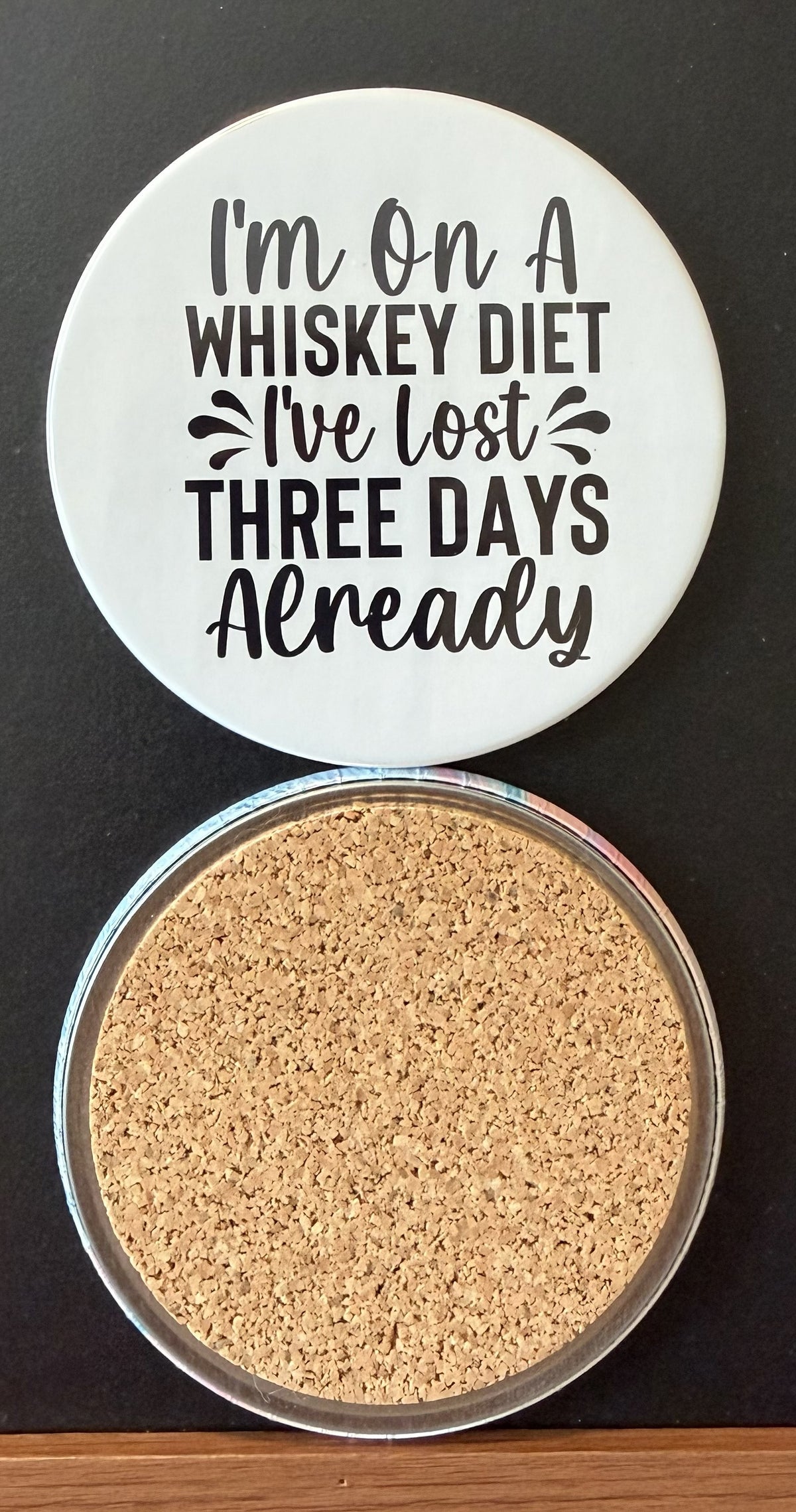 White round drink coaster with a cork backing with black DECORATIVE ACCENTS with text 'I'M ON A WHISKEY DIET, I'VE LOST THREE DAYS ALREADY' on a white background.  This photo was taken on a black background.