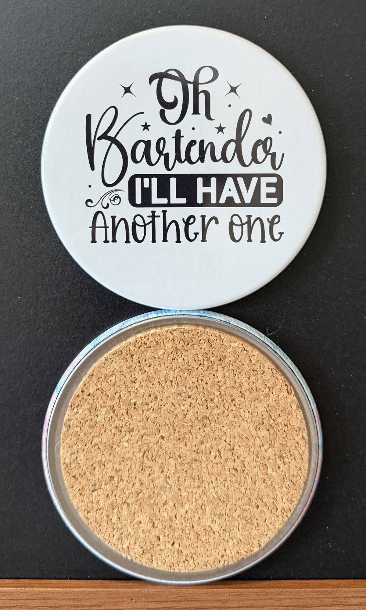 White round drink coaster with a cork backing with black decorative accents with text 'oh bartender, I'll have another one' on a white background.  This photo was taken on a black background.