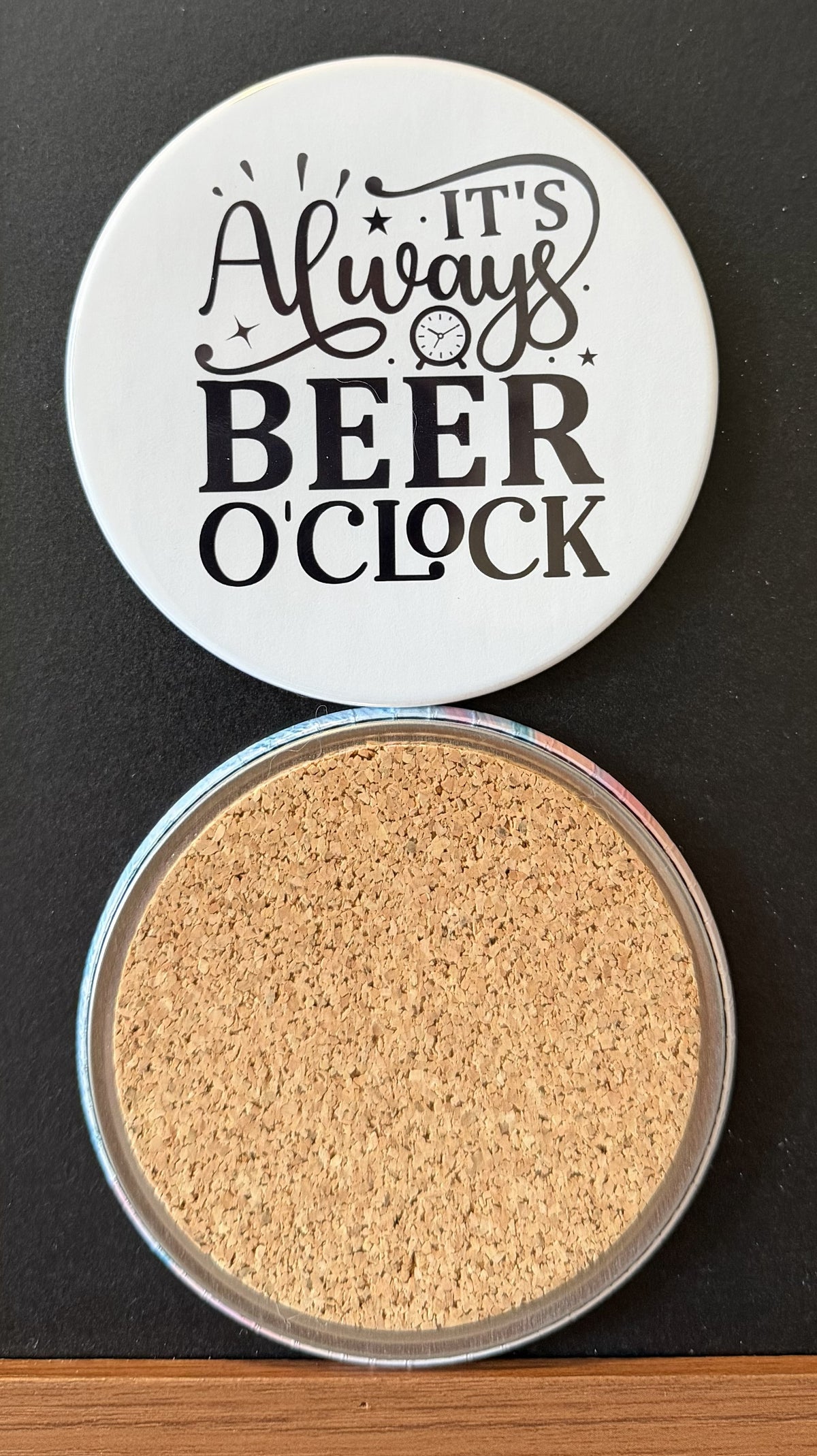 White round drink coaster with a cork backing with a black image of a clock with text 'it's always beer o'clock' on a white background.  This photo was taken on a black background.