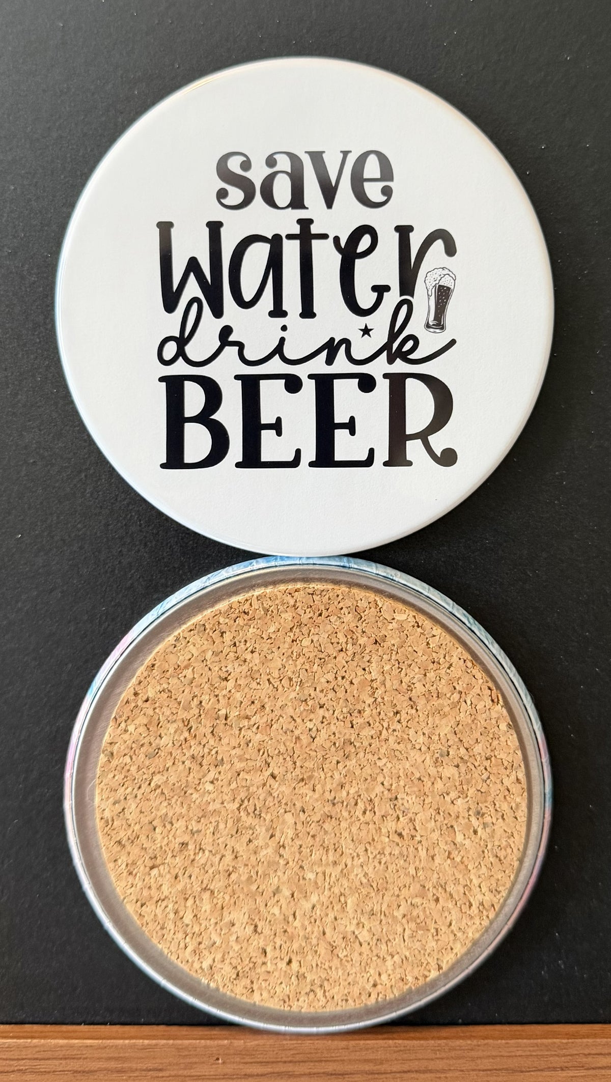 White round drink coaster with a cork backing with a black image of a beer glass with text 'save water, drink beer' on a white background.  This photo was taken on a black background.