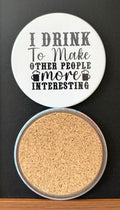 White round drink coaster with a cork backing with 2 black images of beer mugs with text 'i drink to make other people more interesting' on a white background.  This photo was taken on a black background.