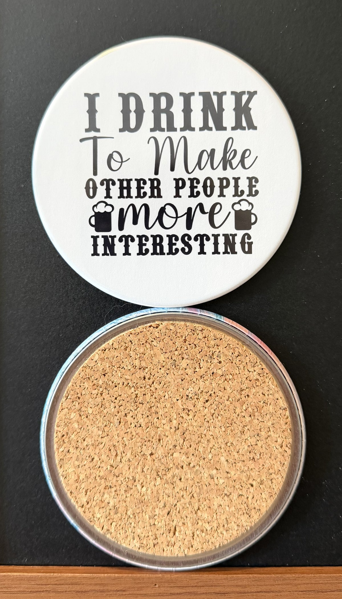 White round drink coaster with a cork backing with 2 black images of beer mugs with text 'i drink to make other people more interesting' on a white background.  This photo was taken on a black background.