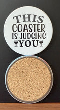 White round drink coaster with a cork backing with 2 black images of cocktail glasses with text 'this coaster is judging you' on a white background.  This photo was taken on a black background.