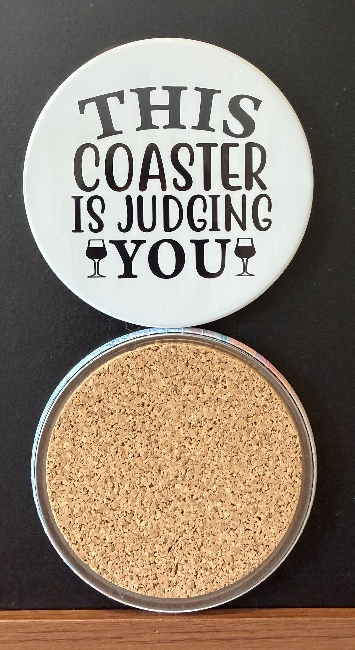 White round drink coaster with a cork backing with 2 black images of cocktail glasses with text 'this coaster is judging you' on a white background.  This photo was taken on a black background.