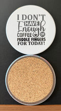 White round drink coaster with a cork backing with a black image of a steaming cup of coffee with text 'I don't have enough coffee or middle fingers for today' on a white background.  This photo was taken on a black background.