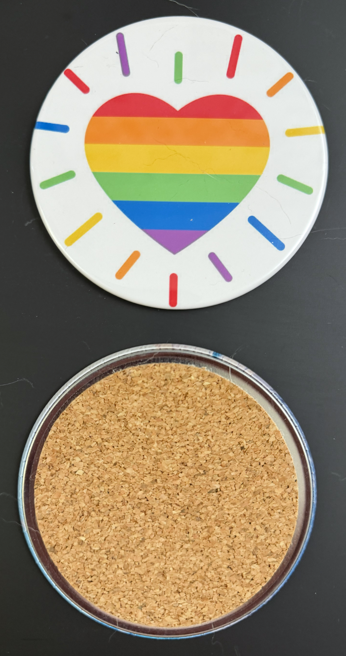 White round drink coaster with a radiating rainbow heart image, with a cork backing  This photo was taken on a black background.
