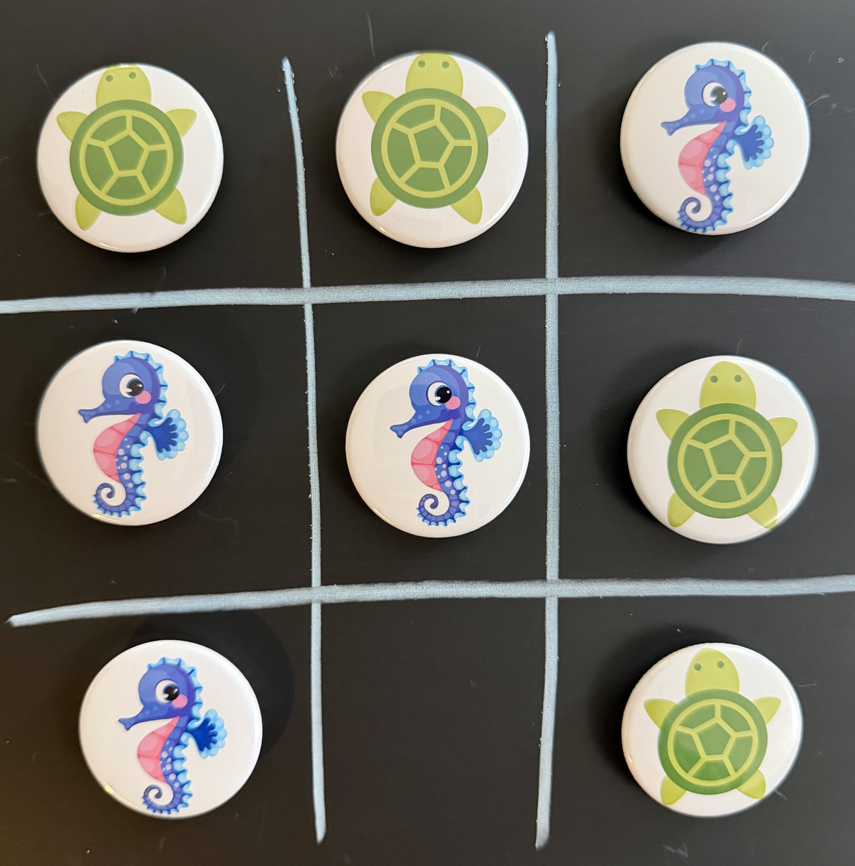 8 Tic Tac Toe small round magnets on a black background with a white grid pattern.  4 game pieces have  green turtle images and the other 4 images are pink and purple seahorses.