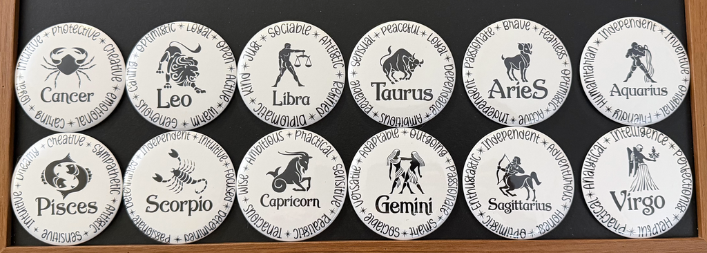 Set of 12 white zodiac round magnets / buttons with black text of the 12 zodiac signs with each button displaying different characteristics associated with each zodiac sign, on a dark background.