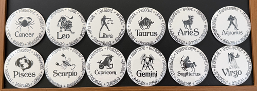 Set of 12 white zodiac round magnets / buttons with black text of the 12 zodiac signs with each button displaying different characteristics associated with each zodiac sign, on a dark background.