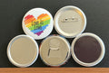 Round white magnet with a rainbow colored heart image with 'Love is Love' in black text.  Also in the photo are the four options available for this magnet which include a pin back button, pocket mirror, magnetic bottle opener and magnet.  This photo was photographed on a black background.