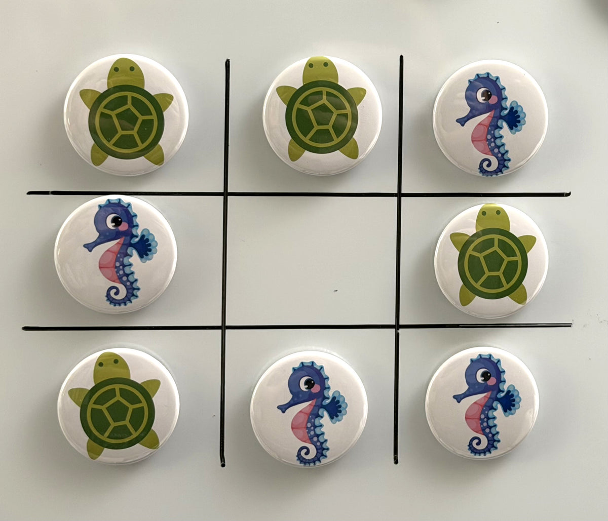 8 Tic Tac Toe small round magnets on a white background with a black grid pattern.  4 game pieces have  green turtle images and the other 4 images are pink and purple seahorses.
