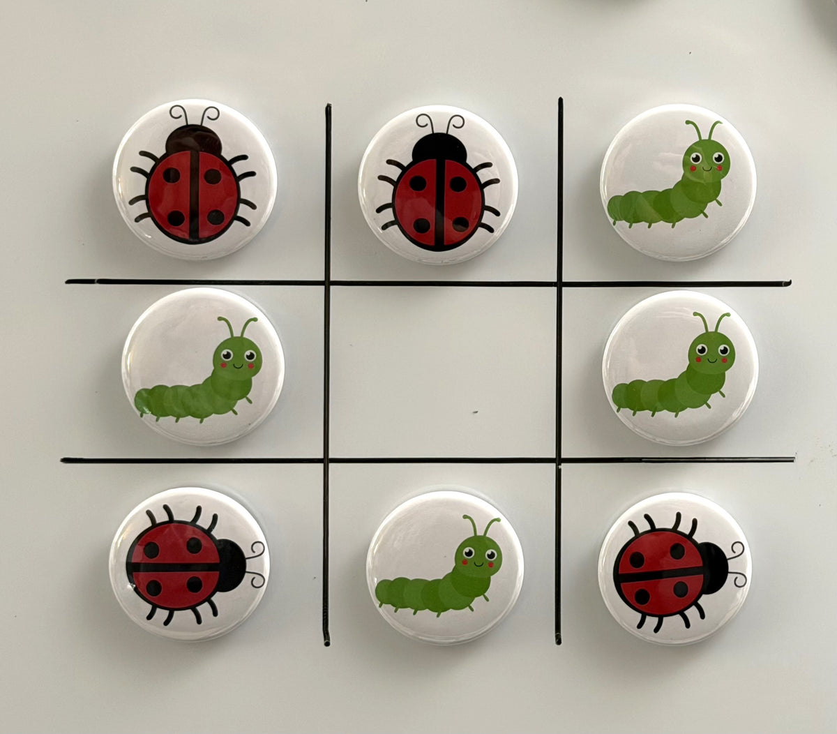 8 Tic Tac Toe small round magnets on a white background with a black grid pattern.  4 game pieces have  red and black lady bug images and the other 4 images are green caterpillars with a smiling face.  