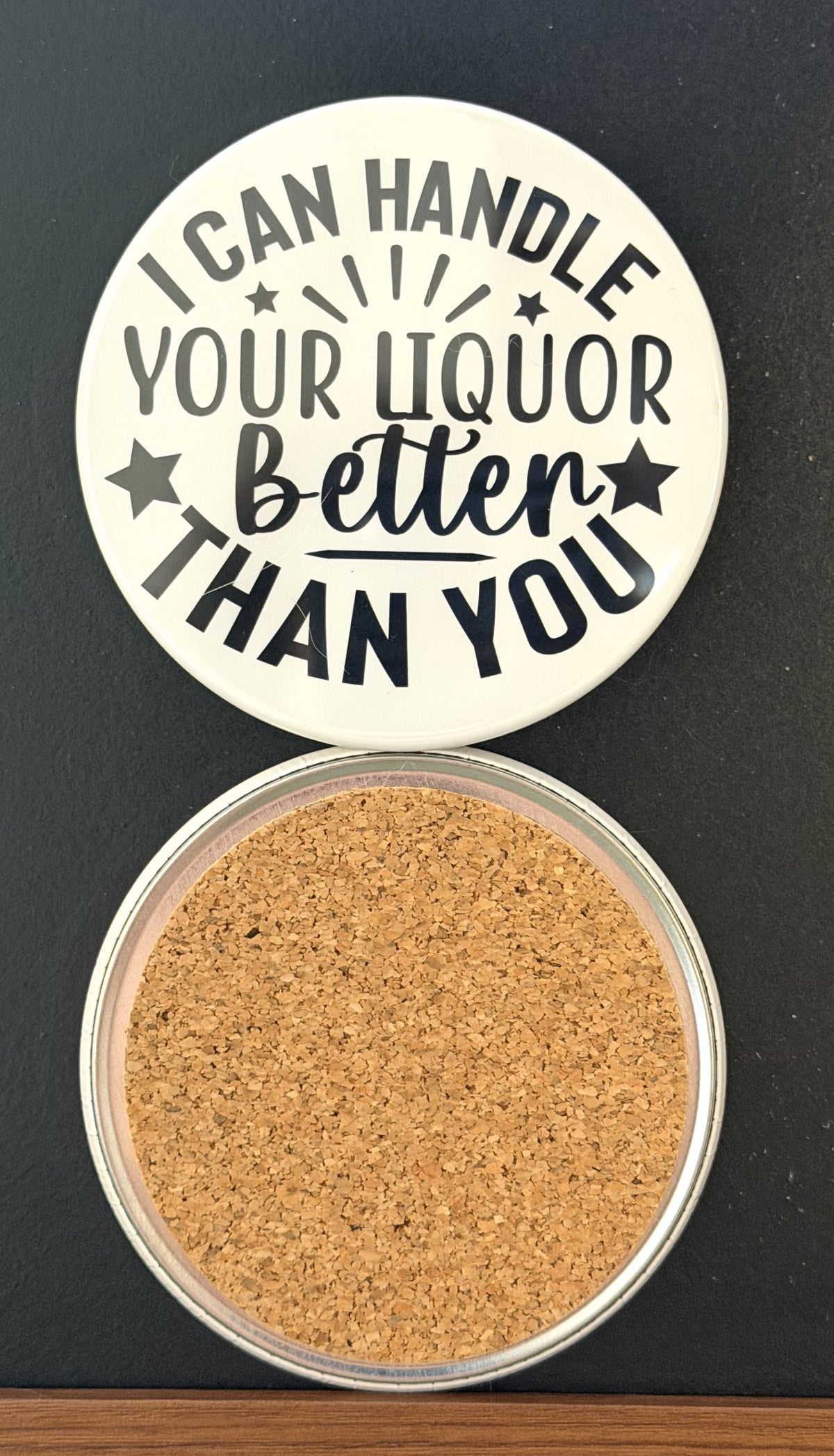 Round white drink coaster with cork backing with 'I can handle your liquor better than you' in black text, with black stars, photographed on a black background.