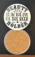 Round white drink coaster with cork backing with 'Beauty is in the eye of the beer holder' in black text, a black arrow and heart, photographed on a black background.