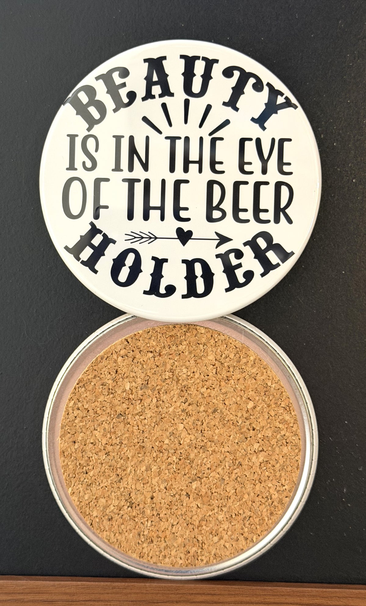 Round white drink coaster with cork backing with 'Beauty is in the eye of the beer holder' in black text, a black arrow and heart, photographed on a black background.