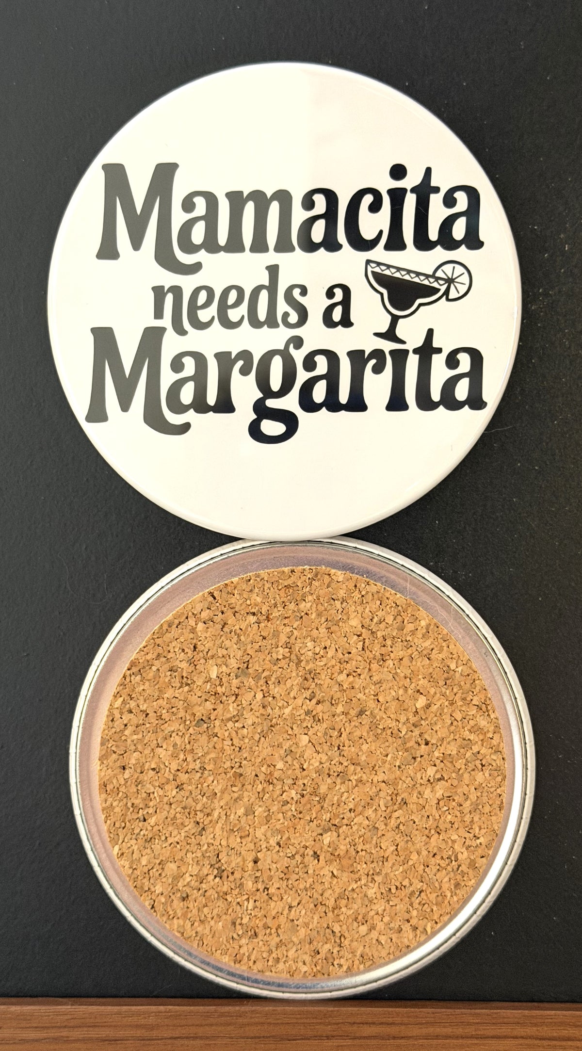 Round white drink coaster with cork backing with 'Mamacita needs a margarita' in black text, with an image of a black margarita glass, photographed on a black background.