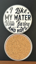 Round white drink coaster with cork backing with 'I like my water with barley and hops' in black text, with an image of a black margarita glass, photographed on a black background.
