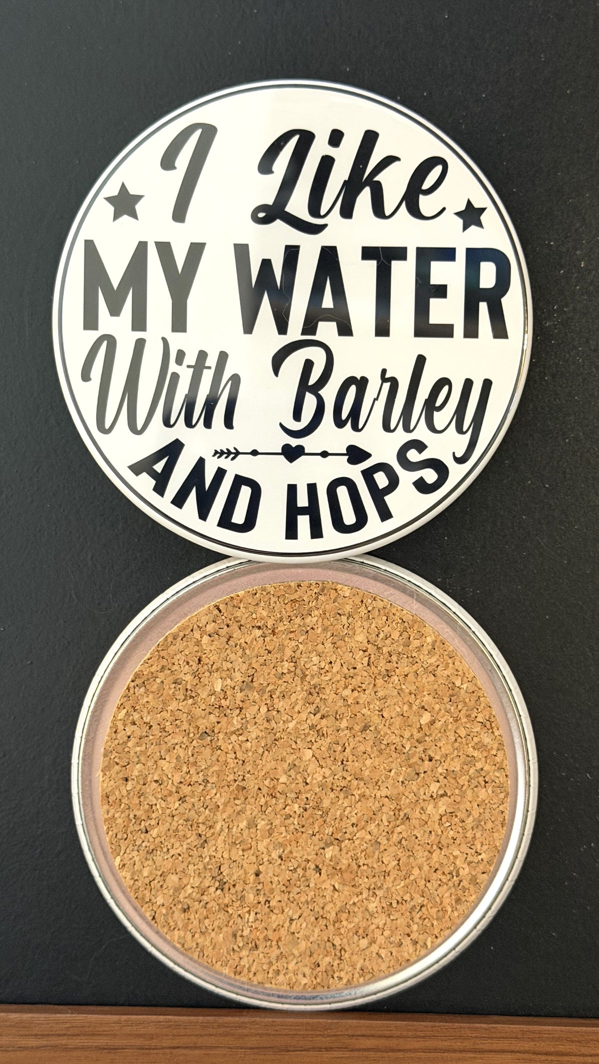 Round white drink coaster with cork backing with 'I like my water with barley and hops' in black text, with an image of a black margarita glass, photographed on a black background.