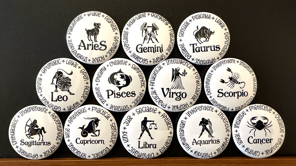 Set of white round magnets/buttons with black text each of the 12 zodiac signs with each button displaying different characteristics associated with each zodiac sign, arranged in a pyramid formation on a dark background.