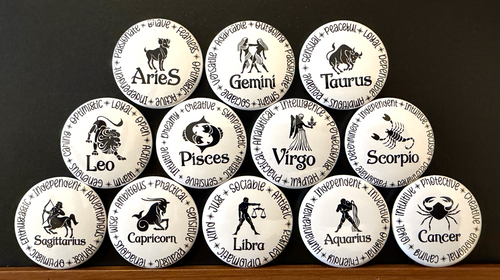 Set of white round magnets/buttons with black text each of the 12 zodiac signs with each button displaying different characteristics associated with each zodiac sign, arranged in a pyramid formation on a dark background.