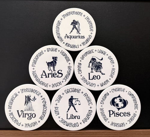 Set of 6 white zodiac drink coasters with dark navy blue text each of the 6 of 12 zodiac signs with each button displaying different characteristics associated with each zodiac sign, arranged in a pyramid formation on a dark background.