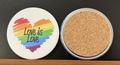 White round drink coaster with a cork backing with a rainbow heart image and 'Love is Love' in black text.  This photo was taken on a black background.