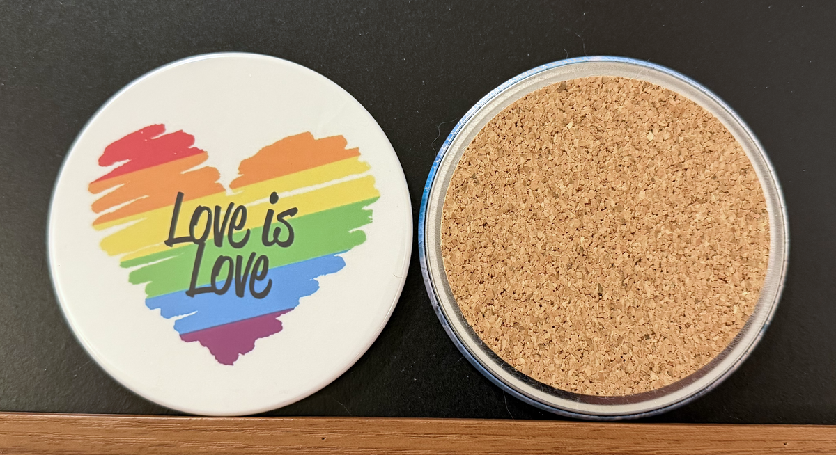White round drink coaster with a cork backing with a rainbow heart image and 'Love is Love' in black text.  This photo was taken on a black background.