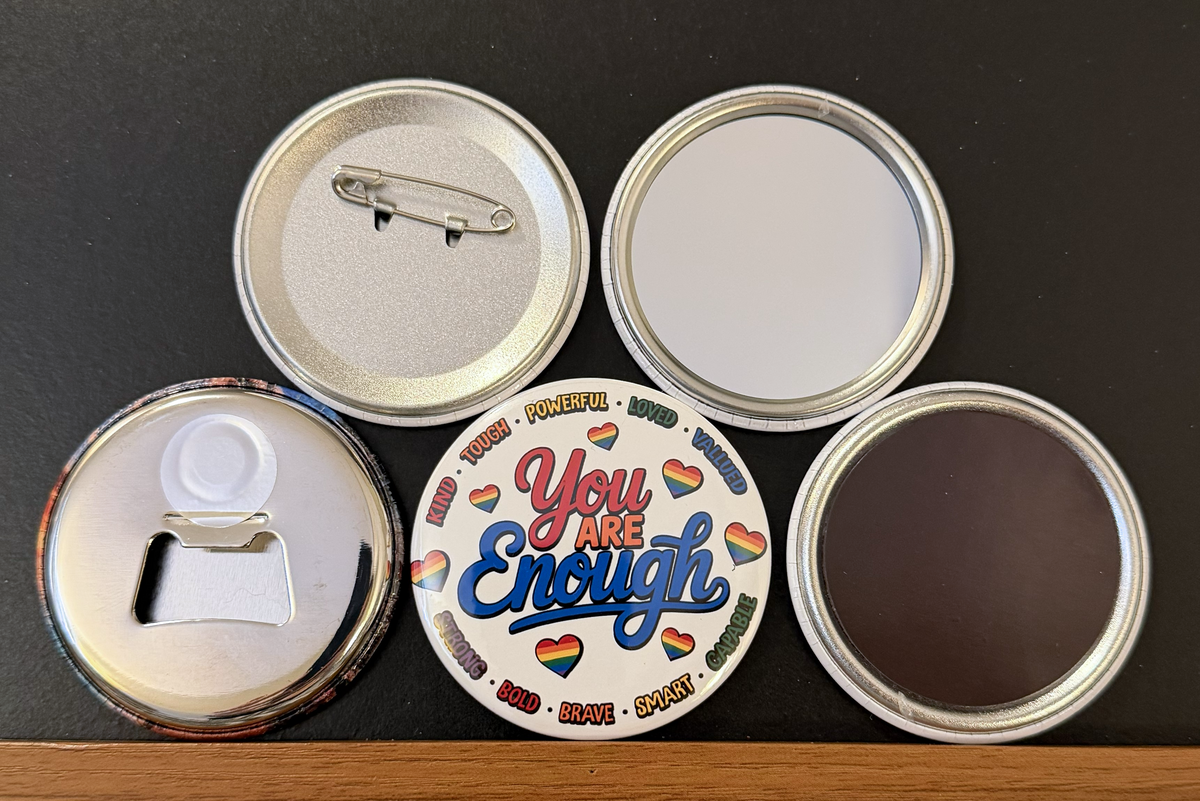 Round white magnet with the message 'you are enough' in red, orange and blue text.  multicolored heart images along with multi-colored text for words such as powerful, loved, valued, smart, bold etc... around the edges of the round magnet/pin.  Also in the photo are the four options available for this magnet which include a pin back button, pocket mirror, magnetic bottle opener and magnet.  This photo was photographed on a black background.